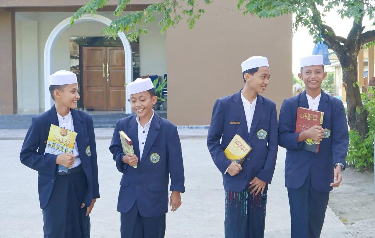 Empat santri berjas biru dongker dan berpeci putih tersenyum sambil membawa kitab di halaman pondok.